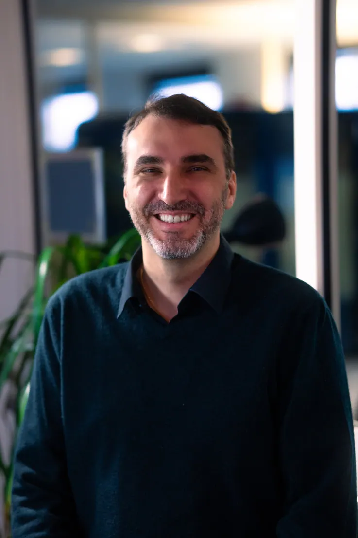 Portrait corporate assis près d'une baie vitrée, en lumière naturelle : collaborateur souriant en chemise sombre